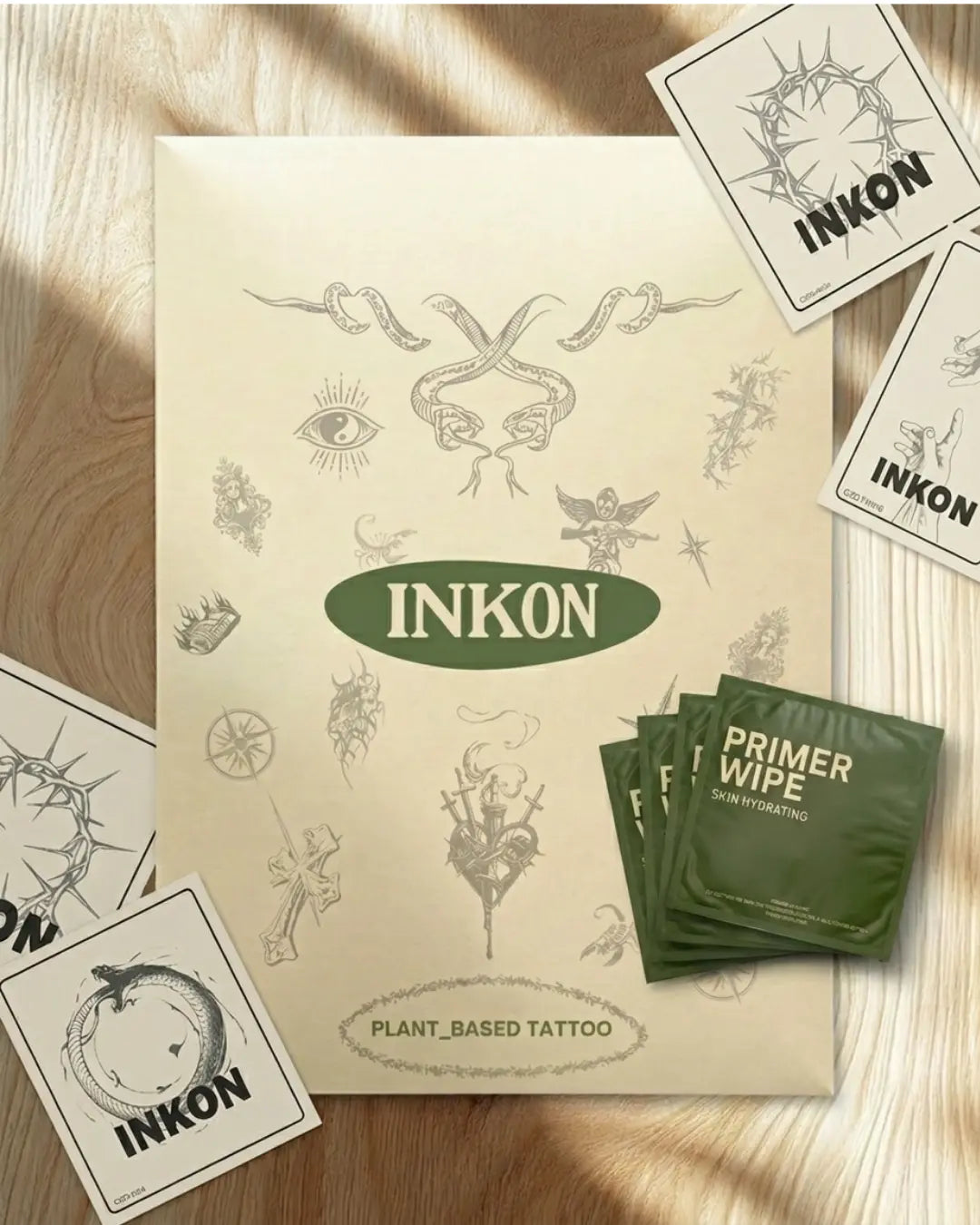 INKON Tattoos For Now complete packaging kit on marble surface – green branded envelope with floral and butterfly illustrations, skin hydrating primer wipe sachet, how to apply instruction card with QR code, and INKON Tattoo branded sticker